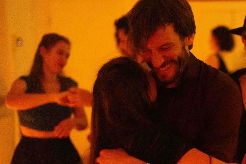 Un homme barbu et souriant tient sa partenaire contre lui pendant une danse blues, dans une lumière orangée