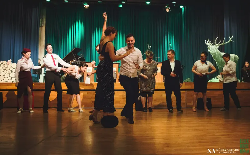 Weitwinkelblick in einen Festival-Tanzsaal mit Paaren auf Holzboden und einer Bühne mit grünem Vorhang und Hirschfigur