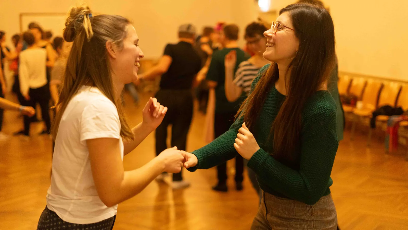 Zwei lachende Frauen tanzen Hand in Hand in einem golden beleuchteten Tanzsaal voller Tänzer
